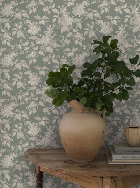 This Syren wallpaper in green features a delicate silhouette pattern of lilac branches and small roses, surface printed in beige and cream on a powdery jade background.